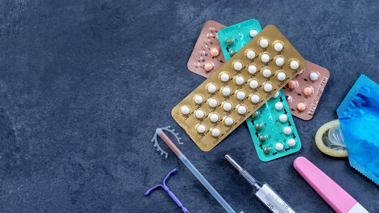 Contraceptive pill packets, an intrauterine device, an implant applicator and a condom laid out on a dark surface.