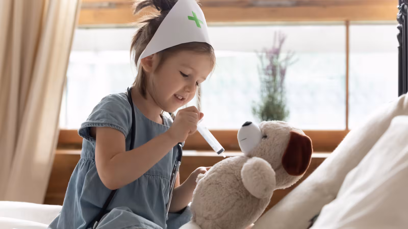 A young child wearing a paper nurse's hat, playing at examining a soft toy dog with a toy thermometer.
